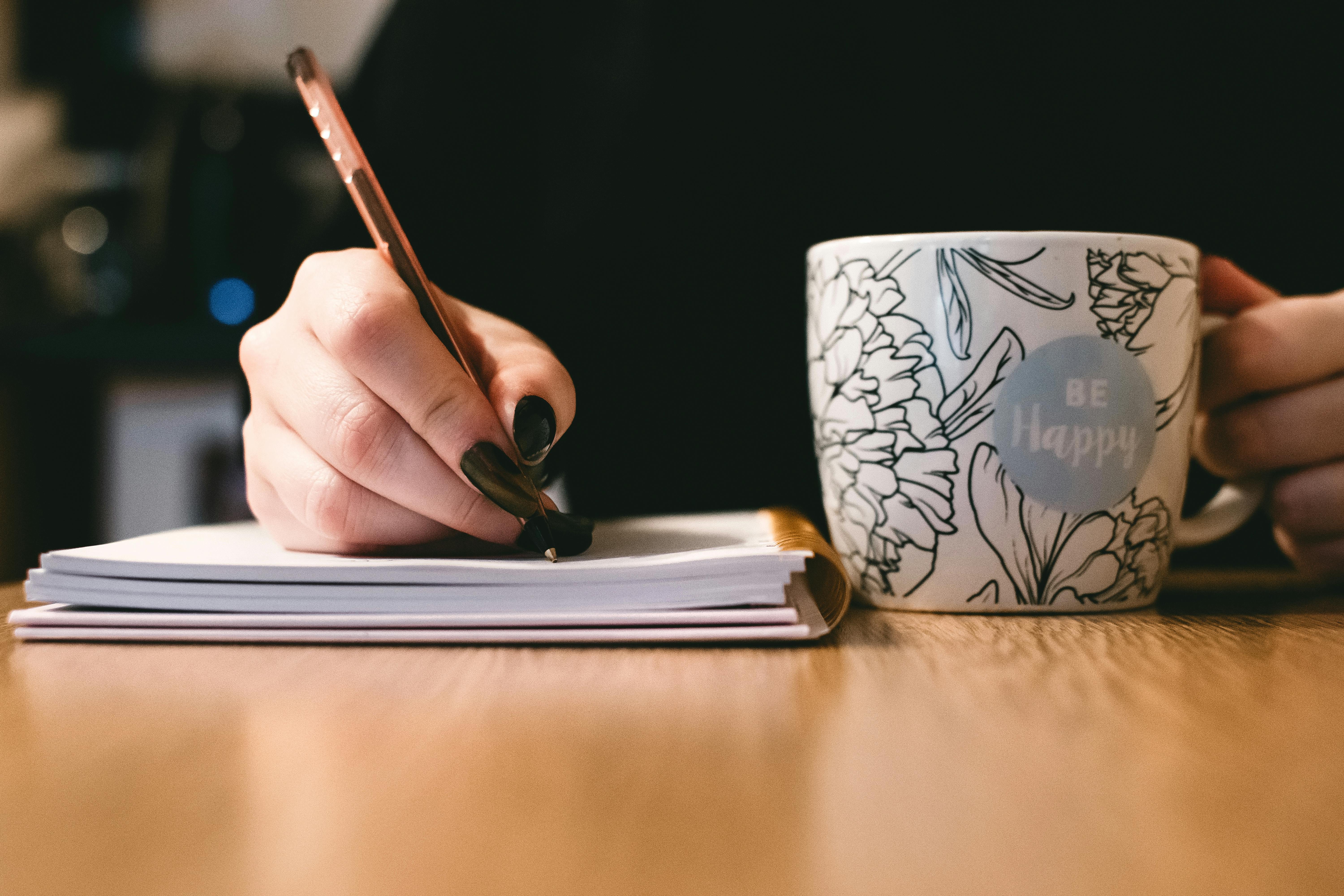 Autorin schreibt auf einen Block, daneben steht eine Tasse mit der Aufschrift 'be happy'
